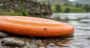 A highly detailed, photorealistic close-up photograph capturing the macro textures of the specific orange rubber 'AQUAFLIGHT' frisbee. The frisbee is resting partially submerged at the water's edge of a natural British lake, illuminated by soft natural daylight. The focus is critically sharp on a single, visible puncture mark and a superficial scratch on the durable, textured natural rubber surface, demonstrating its 'toughness' after use. Droplets of clear water bead on the surface and the surrounding wet, dark sand and smooth river stones.