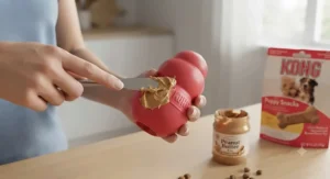 A red rubber KONG puppy toy being stuffed with peanut butter using a small spatula in a kitchen.